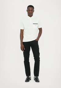 Polo alb cu mânecă scurtă, cu detaliu albastru închis la buzunar, asociat cu pantaloni negri și încălțăminte neagră; textură netedă, design clasic al gulerului.