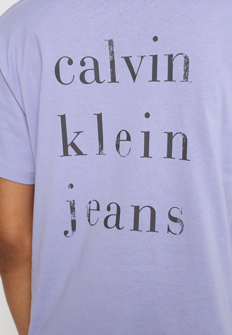 Lawendowa koszulka z czarnym, postarzanym napisem "calvin klein jeans" nadrukowanym w pionie na plecach.