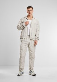 Lichtbeige sportjacket met elastische mouwen en bijpassende broek. Glad materiaal, ritssluiting en zijzakken. Casual trainingsschoenen.