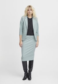 Svetlo zelena krojena blazer in ujemajoča se svila krila, v kombinaciji s črno majico in črnimi čevlji do kolen; gladka tkanina in prilegan kroj.