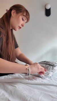 Jeune femme aux longs cheveux ajuste ou plie une couverture à motifs en étant assise sur un lit dans une chambre minimaliste.