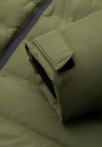 Gros plan sur une manche de veste matelassée vert olive avec une sangle velcro réglable et un détail de poignet intérieur noir.
