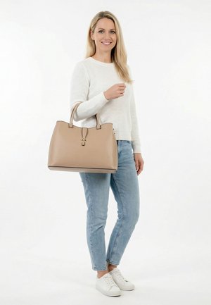 Frau in weißem Pullover und Jeans, die eine große beige Handtasche hält, steht lächelnd vor einem weißen Hintergrund.