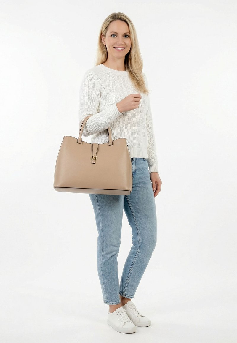 Frau in weißem Pullover und Jeans, die eine große beige Handtasche hält, steht lächelnd vor einem weißen Hintergrund.