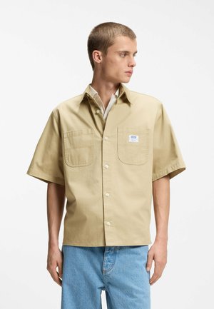 Ung mand iført beige kortærmet skjorte med knapper og to brystlommer samt lyseblå jeans, der ser mod sin venstre side mod en hvid baggrund.