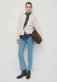 Kobieta w kremowym kardiganie na czarnej koszuli w kratę, jasnoniebieskich dżinsach, czarnych botkach, trzymająca dużą brązową zamszową torbę na tle białej ściany.