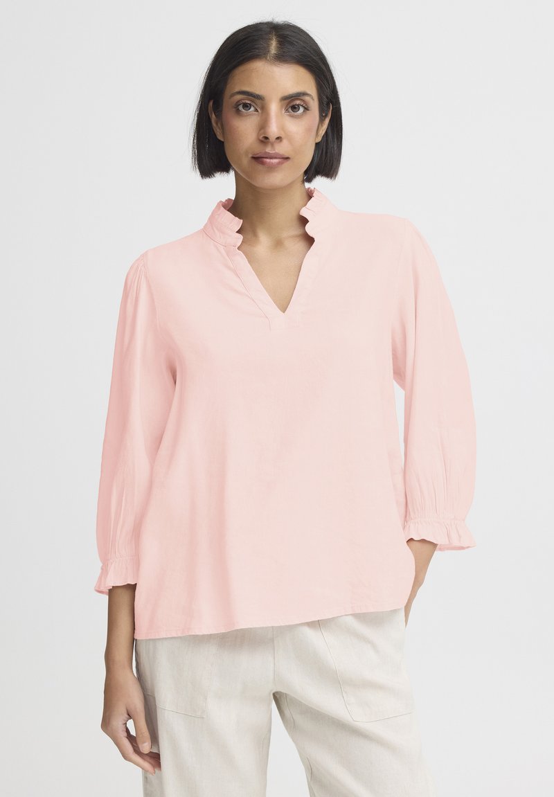 Blouse rose clair avec un col à volants et des manches trois-quarts bouffantes. Confectionnée dans un matériau doux, elle présente un décolleté en V.