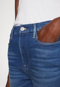 Jeans de mezclilla azul oscuro con un corte ajustado, que cuentan con un bolsillo frontal, botón plateado y un sutil desgaste en los muslos.