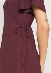 Burgundy wrap dress made of textured fabric, featuring a tie closure at the waist and flutter sleeves for added detail.
