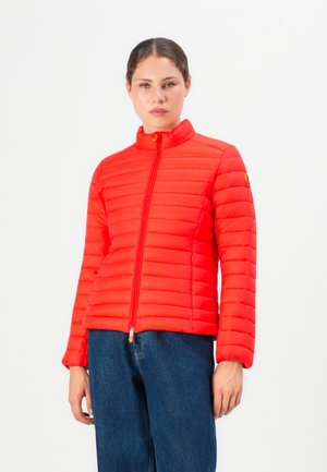 Vrouw in een felrode gewatteerde jas met een ritssluiting aan de voorkant en blauwe jeans, staande tegen een effen witte achtergrond.