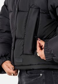 Svart puffjacka med en kortad design, quiltad textur och sidoficka med dragkedja. Har elastiska ärmmuddar och en märkesetikett på framsidan.