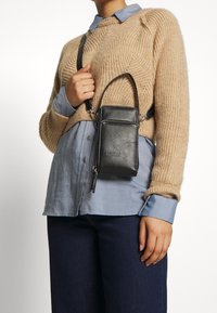 Musta nahkainen olkalaukku, jossa on suorakulmainen muoto, vetoketjukiinnitys ja kohokuvioitu logo. Säädettävä hihna ja sileä pinta.