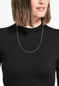 Collana d'oro esposta su una persona che indossa una maglietta nera. La collana presenta un design delicato e intrecciato con una finitura liscia.
