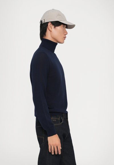 Navyblauer Rollkragenpullover aus glatter Stoff, kombiniert mit dunklen Jeans. Hellbeige Kappe mit Logo, schräg getragen. Seitenansicht.