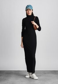 Naine kannab pika musta kõrge kaelusega kleiti, valgeid tossusid, sinist pesapallilotti ja hoiab väikest pruuni õlakotiga lihtsa tausta ees.