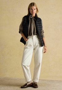 Fekete párnázott mellény kockás ing felett, masnival; fehér csípőszűk nadrág; barna loaferek; textúrált háttér lágy, semleges árnyalatokban.