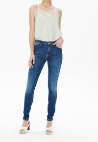 Camisola verde chiaro con pizzo, jeans skinny blu e sandali con tacco bianchi. Il tessuto morbido e il design aderente mettono in risalto la silhouette.