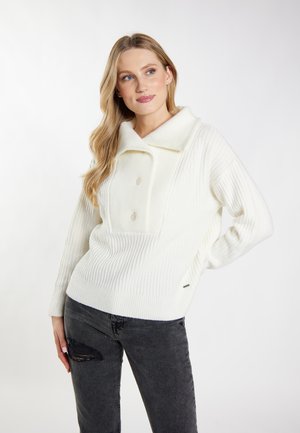 Donna con lunghi capelli biondi che indossa un maglione bianco a bottoni e jeans neri strappati, in piedi con una mano sul fianco.
