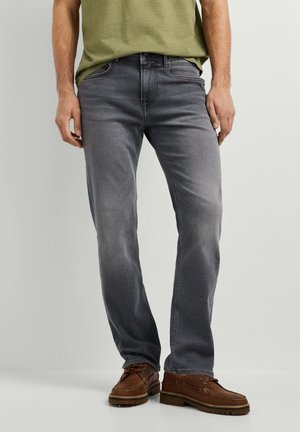 Mann trägt ausgewaschene graue Jeans, braune Lederschuhe und ein olivgrünes T-Shirt und steht vor einem schlichten hellen Hintergrund.