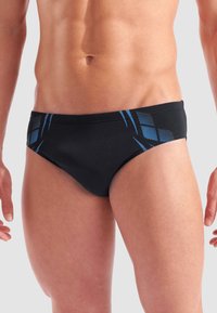 Torso maschile che indossa slip da bagno neri con motivi geometrici blu sui fianchi, in piedi su uno sfondo chiaro e uniforme.