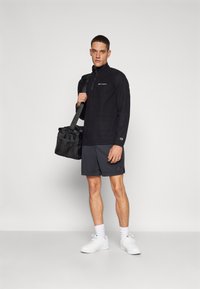 Sudadera de poliéster negro con media cremallera y logo, acompañada de pantalones cortos oscuros. El modelo sostiene un bolso de deporte negro, vistiendo zapatillas blancas y calcetines.