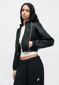 Μαύρο κοντό μπουφάν με λευκές λεπτομέρειες, κατασκευασμένο από ελαφρύ υλικό; συνδυασμένο με λευκό τοπ και μαύρο παντελόνι φόρμας με λογότυπο.