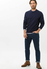 Felpa crewneck navy in cotone, abbinata a jeans blu scuro. Il modello indossa stivali marroni con lacci, in piedi con una postura rilassata.