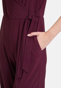 Bordeaux jumpsuit met een strik taille, voorzien van een zijzak en een zachte, rekbare stof. Inclusief geplooid detail bij de taille.