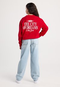 Giovane donna con lunghi capelli ricci in piedi con una mano sulla vita, indossa una felpa rossa con la scritta "La città dei sogni New York" e jeans azzurri chiari.