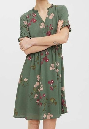 Femme portant une robe midi verte à fleurs avec manches courtes et boutons, bras croisés, montrant des tatouages d'oiseau et d'étoile sur le bras droit.