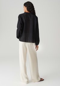 Camicia nera trasparente a maniche lunghe con righe verticali, vestibilità ampia e scollatura rotonda, abbinata a pantaloni a gamba larga color crema.