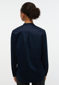 Camicia a maniche lunghe blu navy con collo alto, piega centrale sul retro e polsini con bottone; tessuto liscio con una leggera lucentezza e vestibilità aderente.