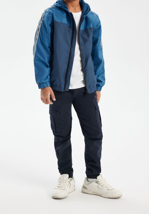 Niño con chaqueta azul con cremallera, camiseta blanca, pantalones cargo azul marino y zapatillas blancas, de pie contra un fondo blanco.