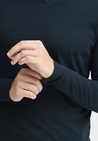 Tamno plava košulja dugih rukava s V izrezom, izrađena od glatkog materijala. Ruke prilagođavaju manžetne, prikazujući prilagođeni dizajn i teksturu košulje.