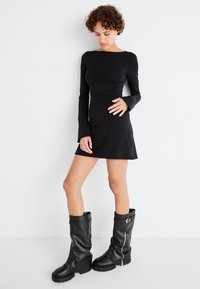 Vestido negro de manga larga con un cuerpo ajustado y dobladillo acampanado, combinado con botas de goma negras hasta la rodilla que cuentan con un zipper lateral y detalle de hebilla.