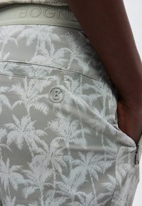 Κοντινό πλάνο γκρι σορτς με λευκό μοτίβο φοίνικα, φερμουάρ πλαϊνής τσέπης και λεπτομέρεια με το λογότυπο «B» κοντά στην τσέπη, φορεμένα από άτομο.