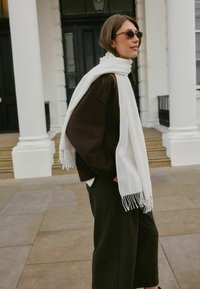 Hvid frynset tørklæde draperet over en mørkebrune sweater. Model iført runde brune solbriller, står på flisebelagte trin foran søjler.