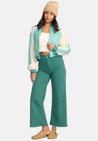 Turquoise and cream bomber jacket with ribbed cuffs, paired with teal high-waisted pants and beige slip-on shoes, featuring a cropped white top.