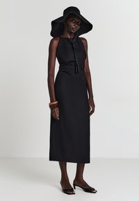 Schwarzes ärmelloses Kleid mit glatter Textur, langen geraden Silhouette und einer Bindung am Hals. Kombiniert mit einem schwarzen Hut mit breiter Krempe.