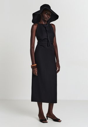 Schwarzes ärmelloses Kleid mit glatter Textur, langen geraden Silhouette und einer Bindung am Hals. Kombiniert mit einem schwarzen Hut mit breiter Krempe.