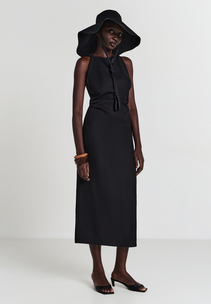 Schwarzes ärmelloses Kleid mit glatter Textur, langen geraden Silhouette und einer Bindung am Hals. Kombiniert mit einem schwarzen Hut mit breiter Krempe.