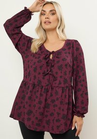 Blusa floreale bordeaux con maniche lunghe, polsini arricciati e nodo frontale. Presenta un tessuto strutturato con vita arricciata per una vestibilità rilassata.
