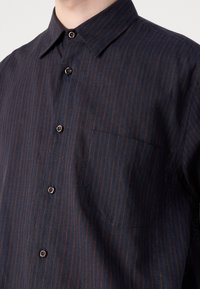 Chemise bleu foncé à boutons avec fines rayures verticales marron et une poche poitrine, portée par une personne avec le bouton du col déboutonné.