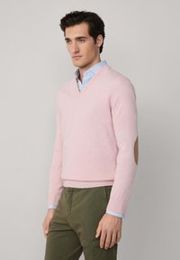 Uomo che indossa un maglione V rosa chiaro con toppe in beige sui gomiti sopra una camicia a righe blu, abbinato a pantaloni verdi oliva, su uno sfondo neutro.