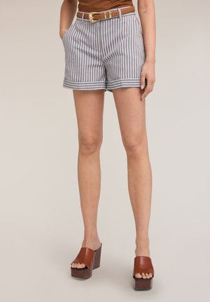 Kobieta ubrana w niebieskie, białe i brązowe pionowo paskowane krótkie spodenki z jasnobrązowym paskiem oraz brązowe sandały na platformie z odkrytymi palcami, stoi z rękami w kieszeniach.