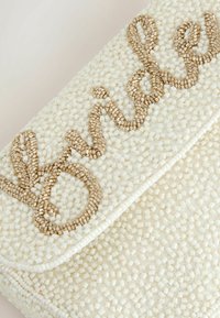 Petite pochette blanche perlée avec le texte cursif "blessed" en perles dorées sur le rabat avant.