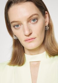 Top verde claro com decote em forma de fechadura. A modelo tem cabelo castanho-claro de comprimento médio e usa brincos circulares prateados com detalhes texturizados.