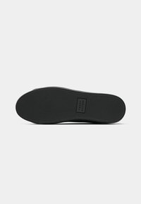 Semelle de chaussure noire avec une texture antidérapante et logo "Tommy Hilfiger" embossé au centre sur un fond blanc uni.