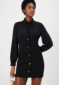 Abito nero con maniche lunghe, colletto e silhouette aderente. Presenta una chiusura frontale con bottoni e due tasche anteriori con accenti in metallo.