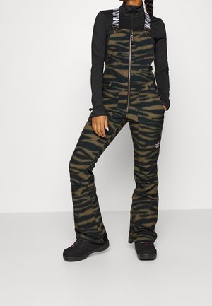 Kobieta ubrana w czarną bluzkę z długim rękawem oraz zielono-czarne ogrodniczki w tygrysie paski zapinane na zamek, z białymi wzorzystymi, regulowanymi ramiączkami i czarnymi butami.